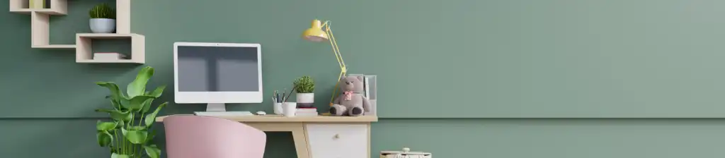 a desk with stationary, a desktop computer, a teddy bear and a lamp, against a green wall