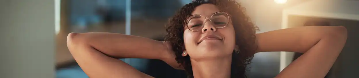 A woman with thick-rimmed clear-framed glasses reclines contentedly in a home office space